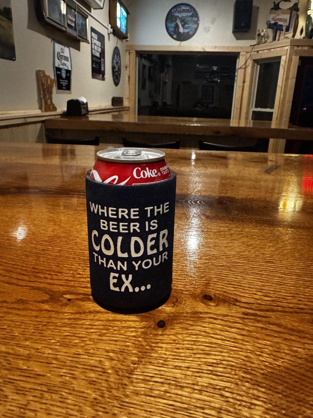 A koozie reading “Where the beer is colder than your ex” on the honey-wood bar top, with the room behind it
